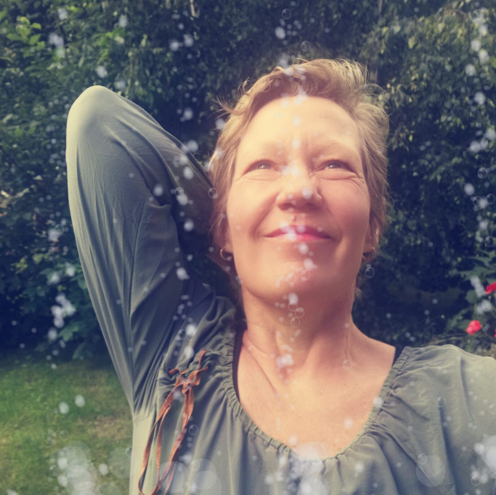 Marion Hanekamp lacht mit geschlossenen Augen im Garten, Wassertropfen und Sonnenlicht um sie herum