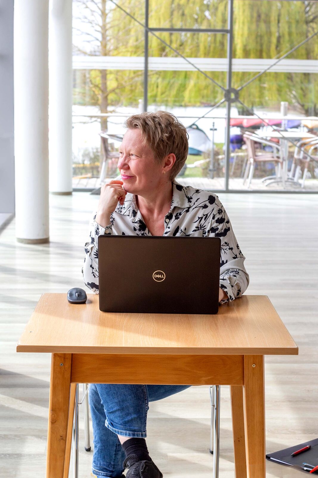 Marion Hanekamp sitzt mit ihrem Laptop an einem Tisch und schaut aus dem Fenster
